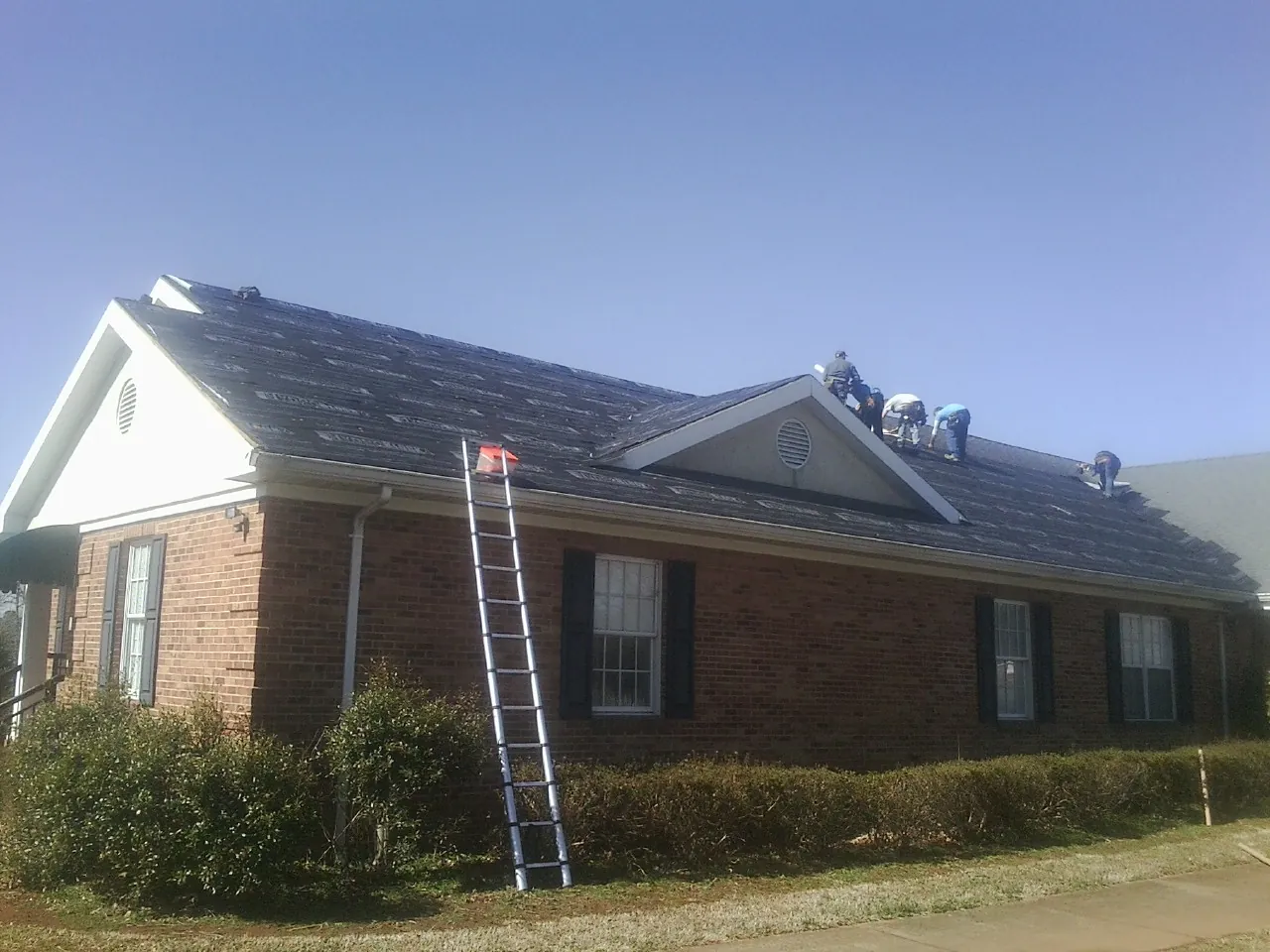 Crew tearing shingles off a brick ranch roof with a ladder set — Metal Roof Repair work by our crew in Brodnax