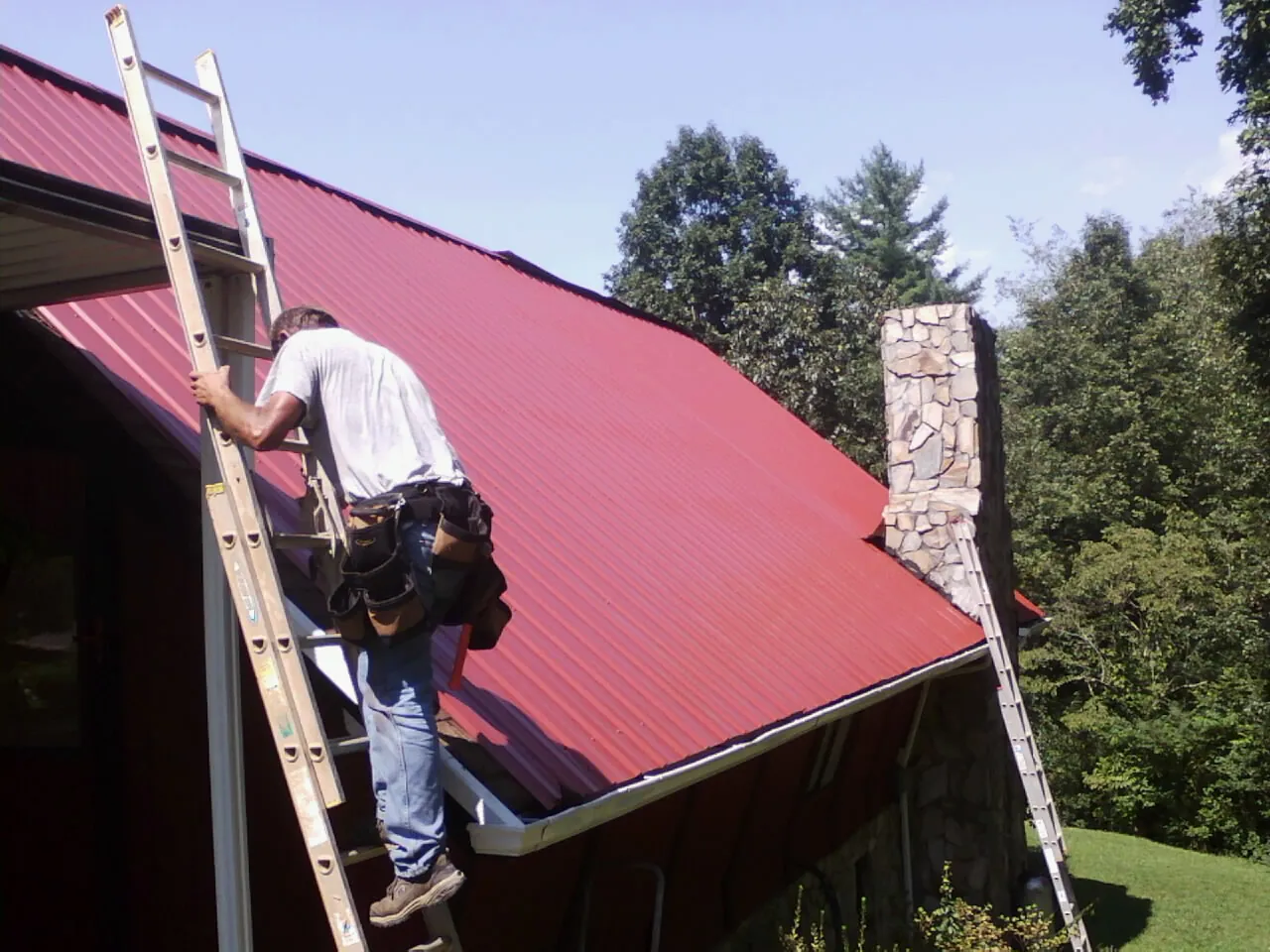 Roofer on a ladder beside a red metal roof and stone chimney — Soffit & Fascia Repair by Roofing Brothers in Brodnax